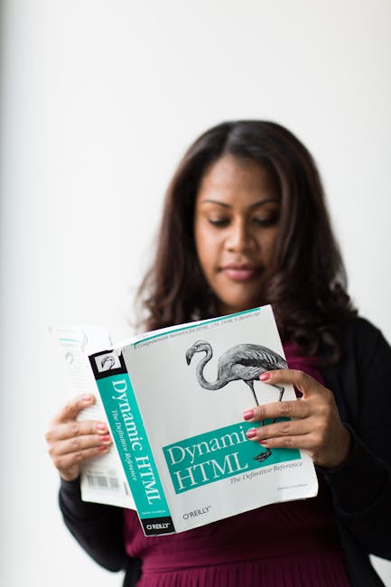 A woman with dark hair, dressed in a black blazer and burgundy top, is standing indoors holding a large book titled 'Dynamic HTML' by O'Reilly. She is looking down at the book, which features an illus